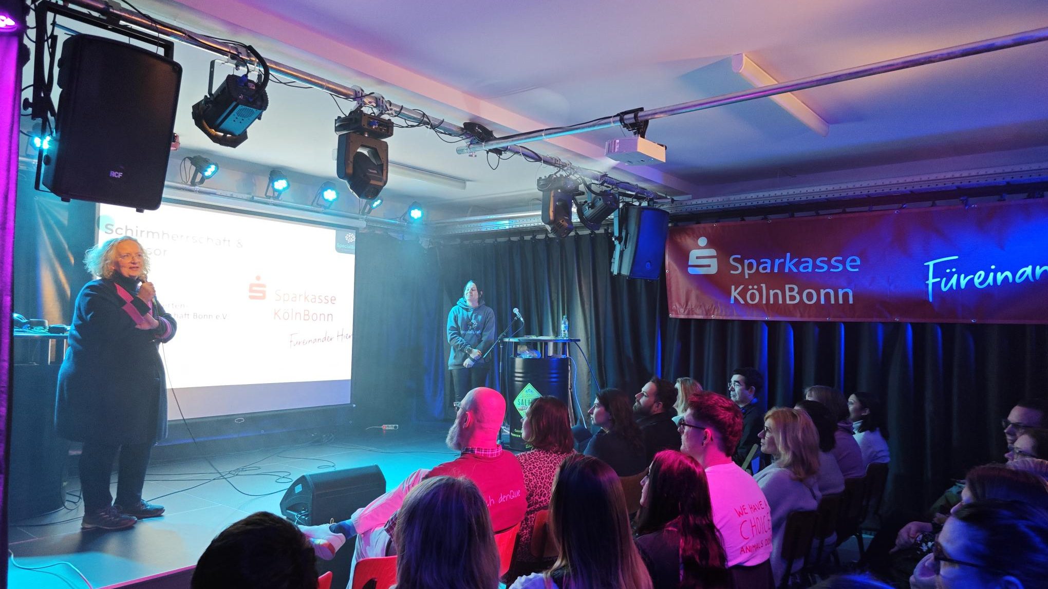auf einer Bühne steht eine Frau mit einem Mikrophon. Sie spricht zu einem Publikum, das vor ihr sitzt.