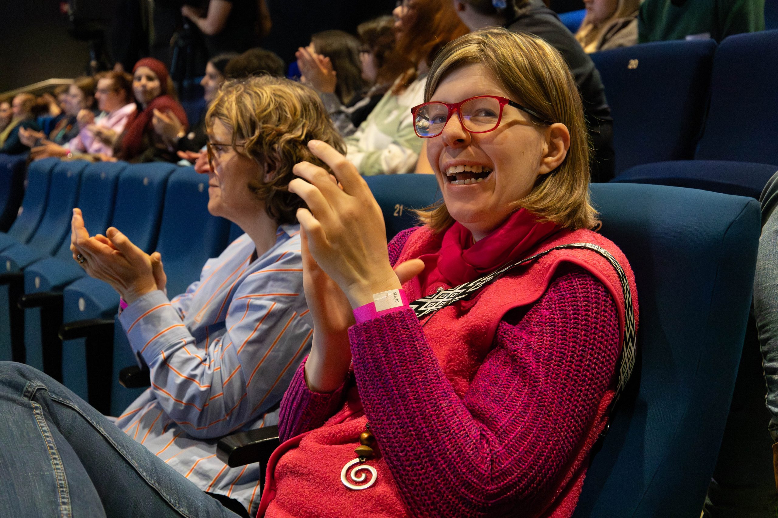 Besucherinnen der Konferenz sitzen im Publikum. Eine Teilnehmerin lacht und klatscht in die Hände.